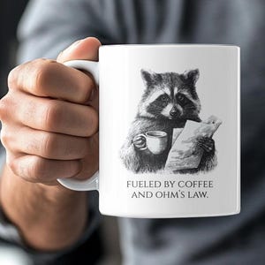 May include: White ceramic mug featuring a black and white illustration of a raccoon holding a coffee cup and a paper. The text on the mug reads "Fueled by Coffee and Ohm's Law."