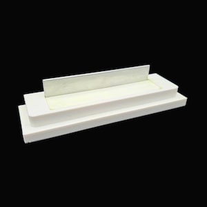 May include: A white, rectangular object with a raised platform and a silver metal blade. The object has a layered design, with a recessed area in the centre. The background is solid black.
