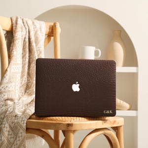 May include: A dark brown laptop cover with a textured crocodile skin pattern and a cutout for the Apple logo. The bottom right corner features the gold-colored embossed initials G&K. The cover is fitted onto a closed laptop.