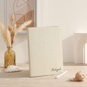 Può includere: Una custodia per tablet color crema con il nome "Aaliyah" in corsivo. La custodia è strutturata e si erge su una superficie chiara. Uno stilo bianco e una conchiglia sono nelle vicinanze. Un vaso con fiori secchi e un'opera d'arte astratta incorniciata sono sullo sfondo.