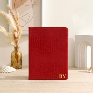 Puede incluir: Un diario rojo texturizado con las iniciales doradas "BY" en la esquina inferior derecha. El diario es el punto focal, colocado sobre un fondo neutro con elementos decorativos como un jarrón y una concha.