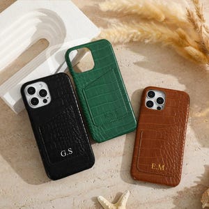 May include: Three phone cases in black, green, and brown, each with a textured crocodile pattern. The black case has the initials "G.S", the green case has "K.S.A", and the brown case has "E.M". The cases have a card slot on the back.