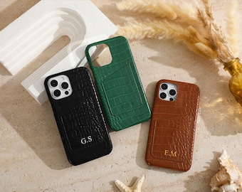 May include: Three phone cases in black, green, and brown, each with a textured crocodile pattern. The black case has the initials "G.S", the green case has "K.S.A", and the brown case has "E.M". The cases have a card slot on the back.