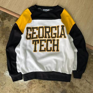 Może przedstawiać: Czarna, biała i złota bluza Georgia Tech. Koszula z długim rękawem ma czarne rękawy i czarny kołnierz. Korpus koszuli jest biały ze złotymi napisami "GEORGIA TECH".