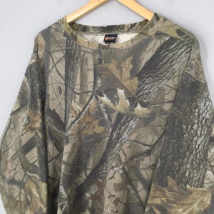 Op de afbeelding: Een sweatshirt met lange mouwen en een ronde hals, voorzien van een realistisch boscamouflagepatroon in tinten bruin, beige en donkergroen. Het kledingstuk heeft een geribbelde halslijn en een klein zwart merklabel in de binnenkraag.