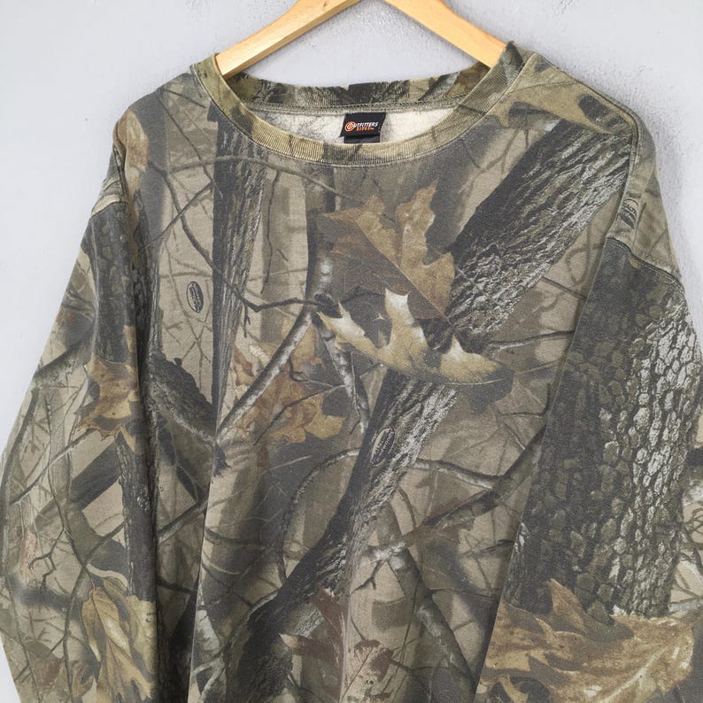 Op de afbeelding: Een sweatshirt met lange mouwen en een ronde hals, voorzien van een realistisch boscamouflagepatroon in tinten bruin, beige en donkergroen. Het kledingstuk heeft een geribbelde halslijn en een klein zwart merklabel in de binnenkraag.