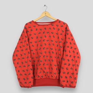May include: A red sweatshirt with a repeating pattern of small cartoon characters. The long-sleeved top has a crew neck and a banded waist and cuffs. The sweatshirt is hanging on a wooden hanger.
