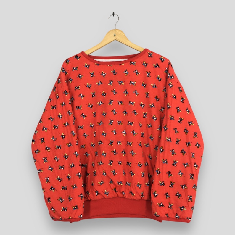 May include: A red sweatshirt with a repeating pattern of small cartoon characters. The long-sleeved top has a crew neck and a banded waist and cuffs. The sweatshirt is hanging on a wooden hanger.