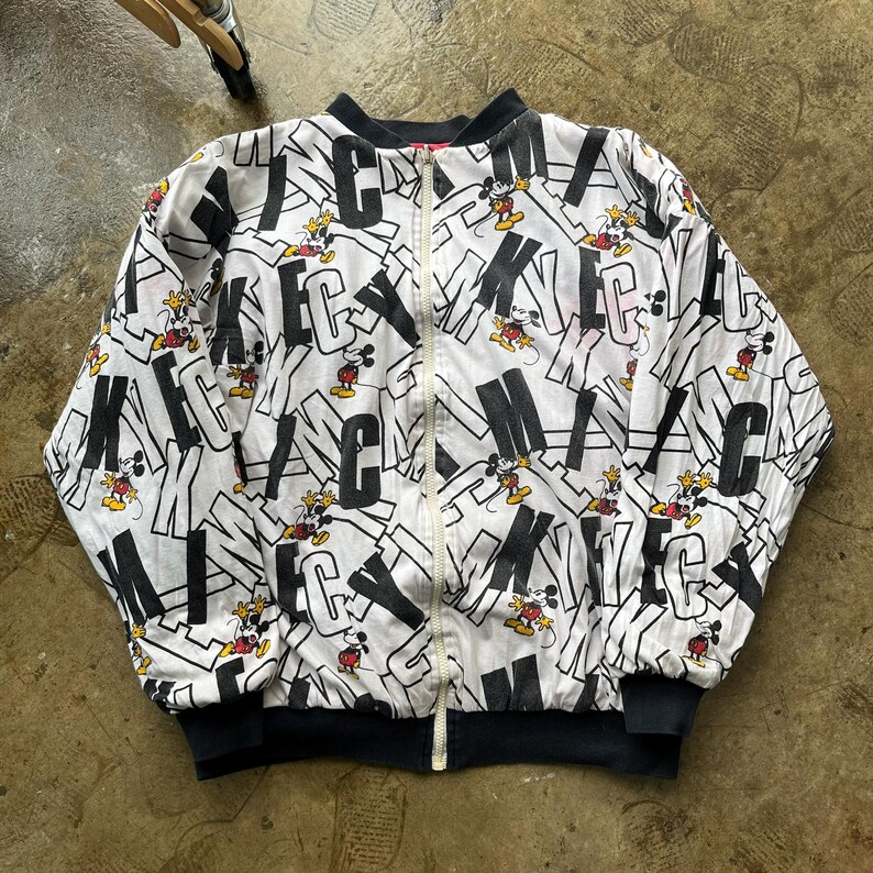 May include: White bomber jacket with a black collar and cuffs. The jacket features a repeating pattern of the word "Mickey" in black block letters, interspersed with cartoon images of Mickey Mouse. The jacket has a full-length zipper.