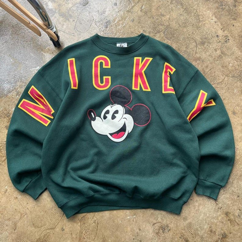 K&ouml;nnte beinhalten: Ein dunkelgr&uuml;nes Sweatshirt mit einem l&auml;chelnden Mickey Mouse-Motiv. Das Wort "Mickey" ist in roten Buchstaben mit gelben Umrissen auf die Brust gedruckt. Die &Auml;rmel tragen ebenfalls die Buchstaben "M" und "Y" im gleichen Stil.