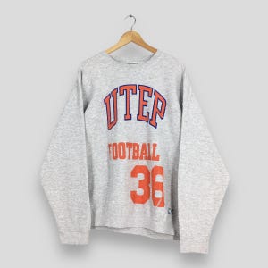 May include: A heather gray sweatshirt with the word "UTEP" in orange and blue lettering. Below the word "UTEP" is the word "FOOTBALL" in orange lettering, and the number "36" in orange. The sweatshirt is hanging on a wooden hanger.