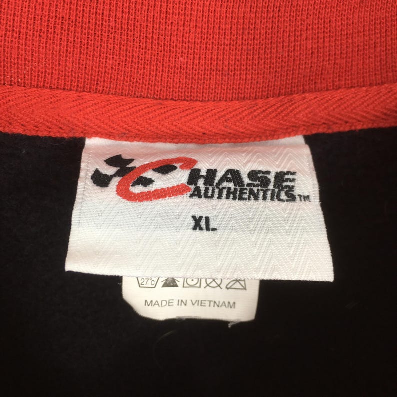 Puede incluir: Primer plano de una prenda negra con cuello rojo. Una etiqueta blanca muestra el texto "CHASE AUTHENTICS" y "XL". La etiqueta tambi&eacute;n incluye instrucciones de lavado y el texto "MADE IN VIETNAM."