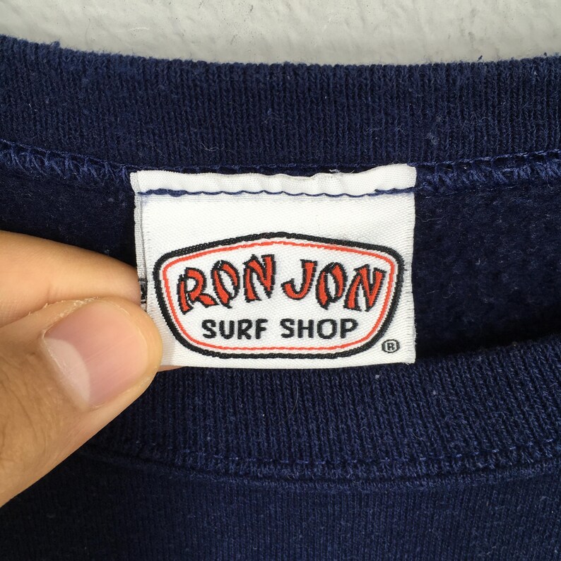 Puede incluir: Primer plano de una etiqueta de sudadera azul marino. La etiqueta presenta las palabras "RON JON SURF SHOP" en rojo y blanco, dentro de un borde ovalado negro. La etiqueta es rectangular con esquinas redondeadas.