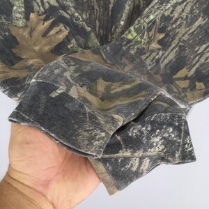 Puede incluir: Primer plano de una camisa de manga larga con estampado de camuflaje. La tela presenta una mezcla de tonos marrones, verdes y grises, creando un dise&ntilde;o de camuflaje de bosque. El pu&ntilde;o de la manga es de color gris oscuro.