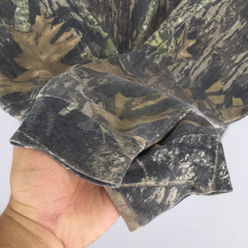 Puede incluir: Primer plano de una camisa de manga larga con estampado de camuflaje. La tela presenta una mezcla de tonos marrones, verdes y grises, creando un dise&ntilde;o de camuflaje de bosque. El pu&ntilde;o de la manga es de color gris oscuro.