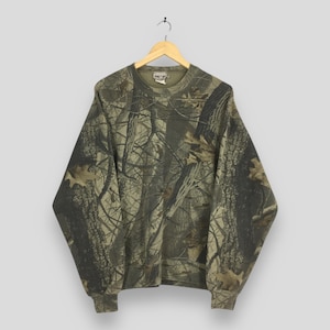 May include: A camouflage-patterned sweatshirt hangs on a wooden hanger. The long-sleeved top features a woodland design in shades of green, brown, and tan. The crew neck sweatshirt has ribbed cuffs and hem.