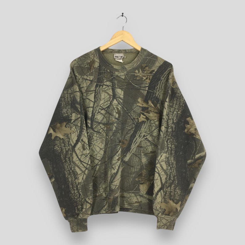 May include: A camouflage-patterned sweatshirt hangs on a wooden hanger. The long-sleeved top features a woodland design in shades of green, brown, and tan. The crew neck sweatshirt has ribbed cuffs and hem.