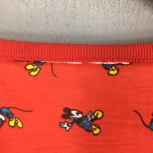 May include: Red fabric with a repeating pattern of Mickey Mouse in various poses. The fabric has a red ribbed collar. The cartoon character is wearing blue shorts, yellow shoes, and white gloves.