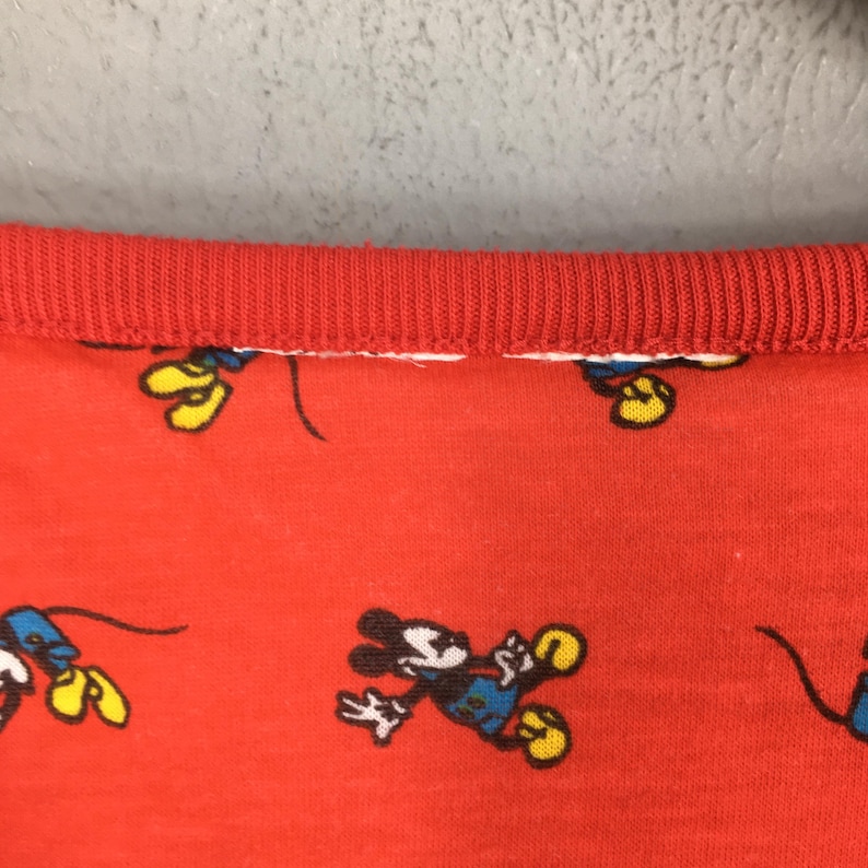 May include: Red fabric with a repeating pattern of Mickey Mouse in various poses. The fabric has a red ribbed collar. The cartoon character is wearing blue shorts, yellow shoes, and white gloves.