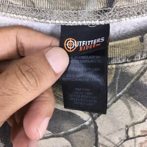 Puede incluir: Primer plano de una etiqueta negra con texto blanco y un logotipo de diana, sujeta a una prenda con estampado de camuflaje. El estampado de la prenda presenta tonos de marr&oacute;n, verde y gris. La etiqueta dice 'Outfitters Ridge'.