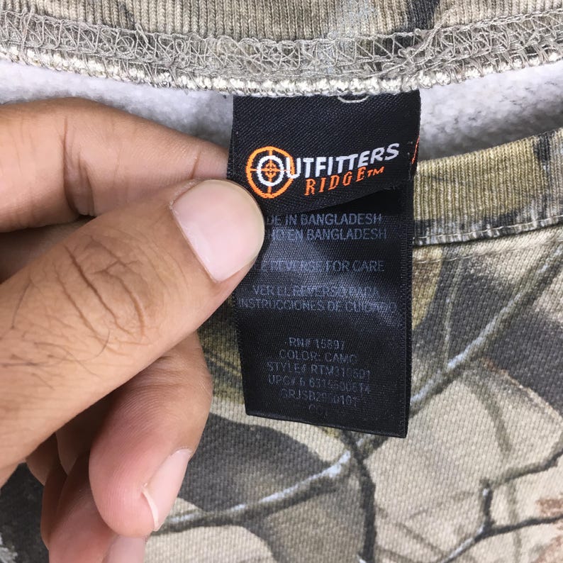 Puede incluir: Primer plano de una etiqueta negra con texto blanco y un logotipo de diana, sujeta a una prenda con estampado de camuflaje. El estampado de la prenda presenta tonos de marr&oacute;n, verde y gris. La etiqueta dice 'Outfitters Ridge'.