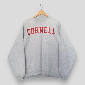 May include: A light gray crewneck sweatshirt featuring the word CORNELL printed in bold red capital letters across the chest. The garment has long sleeves, a ribbed neckline, ribbed cuffs, and a ribbed hem, displayed on a wooden hanger.