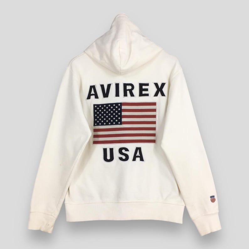 Puede incluir: Sudadera con capucha de color crema con el texto "AVIREX" sobre un parche de la bandera estadounidense y la palabra "USA". La sudadera tiene capucha y mangas largas, con un peque&ntilde;o logotipo en la manga izquierda.