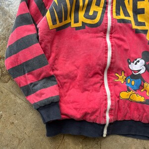 May include: Red zip-up sweatshirt featuring Mickey Mouse. The sweatshirt has black and red striped sleeves and the word "MICKEY" in yellow block letters. Mickey Mouse is on the front, wearing yellow shoes and blue shorts. The sweatshirt has a white zipper and a navy blue hem.