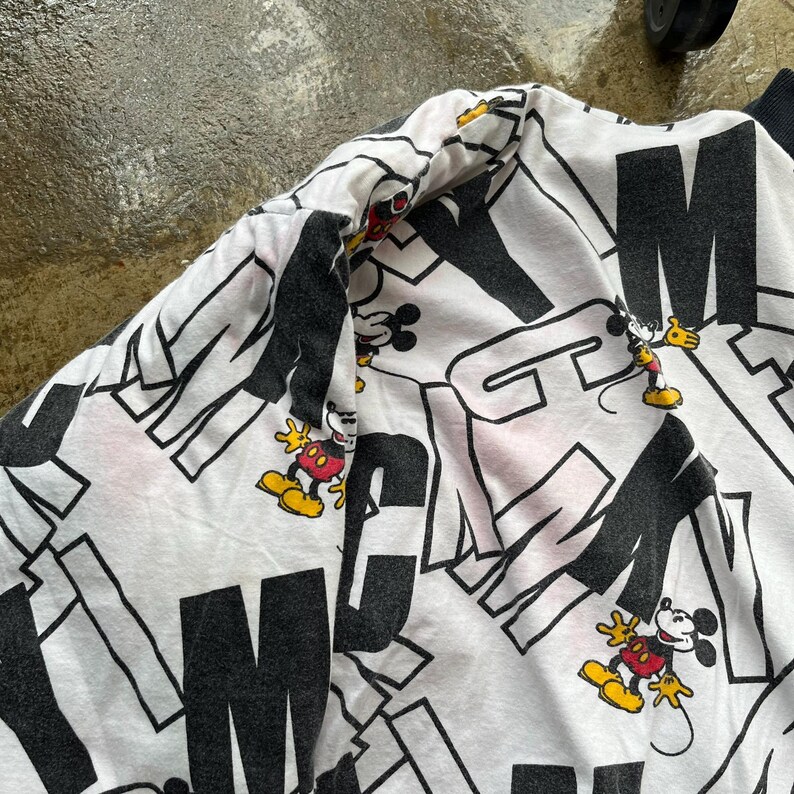 May include: White sweatshirt with a black and white graphic pattern featuring the letters "M" and "N" and cartoon images of Mickey Mouse. The sweatshirt has a black collar and cuffs.