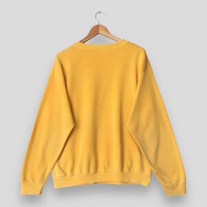 May include: A mustard yellow sweatshirt hanging on a wooden hanger. The crew neck sweatshirt has long sleeves and a ribbed hem and cuffs. The garment is made of a soft, textured material.