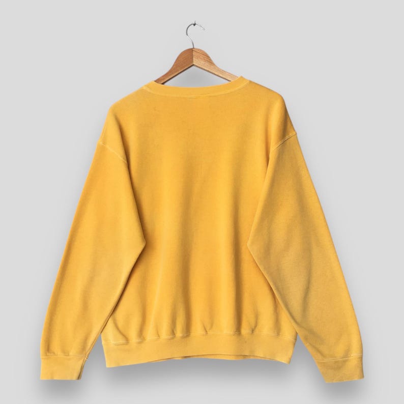 May include: A mustard yellow sweatshirt hanging on a wooden hanger. The crew neck sweatshirt has long sleeves and a ribbed hem and cuffs. The garment is made of a soft, textured material.