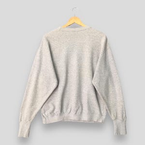 Puede incluir: Una sudadera de cuello redondo en color gris jaspeado claro con mangas largas y pu&ntilde;os acanalados. La prenda presenta un dise&ntilde;o cl&aacute;sico de jersey con cuello y dobladillo acanalados, confeccionada en un tejido suave y texturizado sobre una percha de madera.