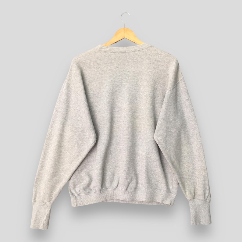Puede incluir: Una sudadera de cuello redondo en color gris jaspeado claro con mangas largas y pu&ntilde;os acanalados. La prenda presenta un dise&ntilde;o cl&aacute;sico de jersey con cuello y dobladillo acanalados, confeccionada en un tejido suave y texturizado sobre una percha de madera.