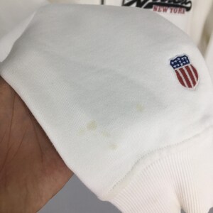 Puede incluir: Sudadera blanca con pu&ntilde;o acanalado y un peque&ntilde;o emblema con un escudo con rayas rojas y un campo de estrellas azules. El texto "NEW YORK" es visible en negro sobre el emblema.