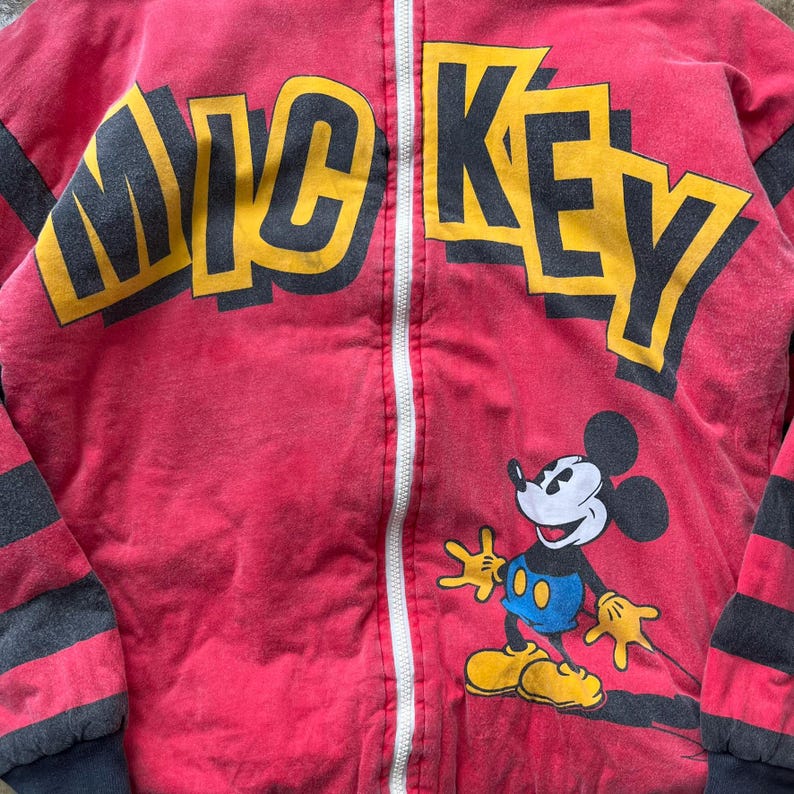 May include: Red zip-up jacket featuring the word "MICKEY" in yellow and black block letters. A cartoon image of Mickey Mouse is on the lower right. The jacket has black and gray striped sleeves.