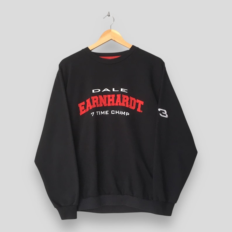 May include: Black crew neck sweatshirt with the text "DALE EARNHARDT 7 TIME CHAMP" in red and white lettering. The number "3" is on the right sleeve in white. The sweatshirt is hanging on a wooden hanger.