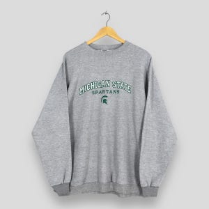 Puede incluir: Sudadera gris de cuello redondo con la inscripción "MICHIGAN STATE SPARTANS" en verde en el pecho. Debajo, un logotipo verde de un casco espartano. La sudadera tiene cuello, puños y dobladillo de canalé.