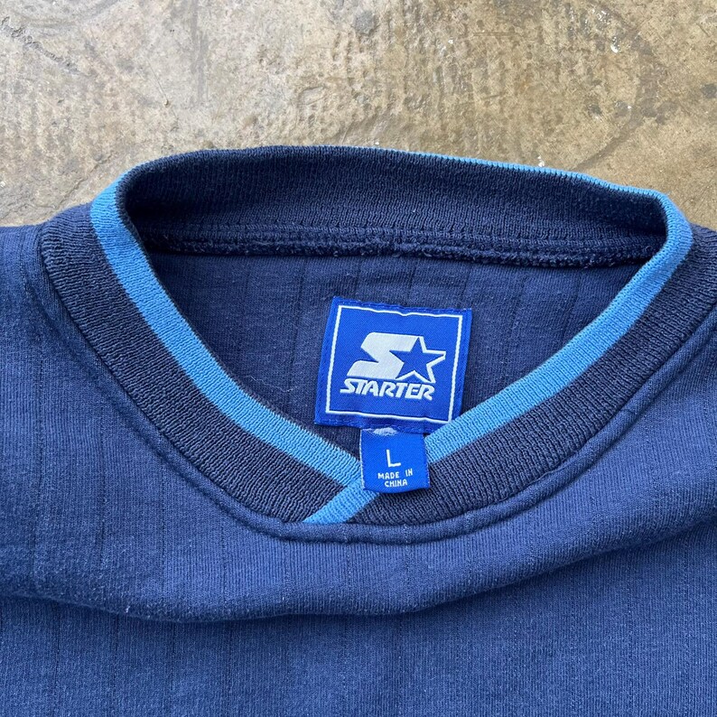 May include: A navy blue long-sleeve shirt with a ribbed texture and a V-neck collar featuring light blue trim. The interior neck label displays the Starter logo and the text L and MADE IN CHINA. The fabric has a consistent dark blue tone.