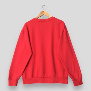 May include: A solid red sweatshirt hangs on a wooden hanger against a light gray background. The crew neck sweatshirt has long sleeves and ribbed cuffs and hem. The back of the sweatshirt is visible, showcasing its simple, clean design.