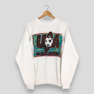 May include: White crewneck sweatshirt featuring a graphic of a soccer ball and the text "USA WORLD SOCCER" in a rectangular design. The graphic has teal, red, and brown accents. The sweatshirt is hanging on a wooden hanger.