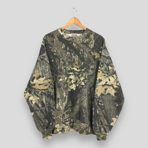 May include: A long-sleeved camouflage sweatshirt hangs on a wooden hanger. The pattern features shades of brown, green, and tan, mimicking a forest environment. The cuffs and neckline are ribbed. The brand name is visible on the neckline.