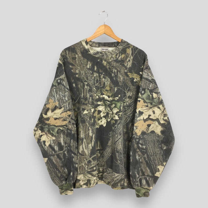 May include: A long-sleeved camouflage sweatshirt hangs on a wooden hanger. The pattern features shades of brown, green, and tan, mimicking a forest environment. The cuffs and neckline are ribbed. The brand name is visible on the neckline.