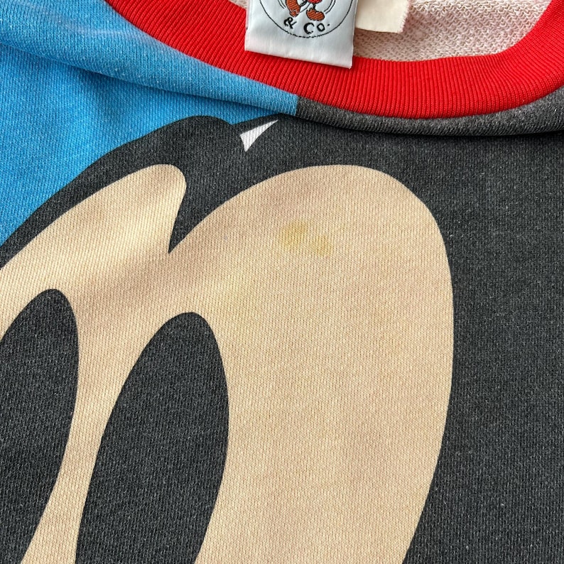 K&ouml;nnte beinhalten: Nahaufnahme eines Vintage-Sweatshirts mit einem gro&szlig;en Mickey Mouse-Gesichtsmotiv. Das Sweatshirt hat ein Schwarz-Beige-Farbschema mit blauen und roten Zierleisten. Auf dem Etikett steht "& Co."