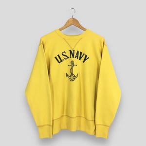 May include: A bright yellow long-sleeve crewneck sweatshirt featuring the text U.S. NAVY in black lettering arched over a black graphic of a ship anchor with a chain. The garment has a ribbed collar, cuffs, and hem with a V-stitch detail at the neck.
