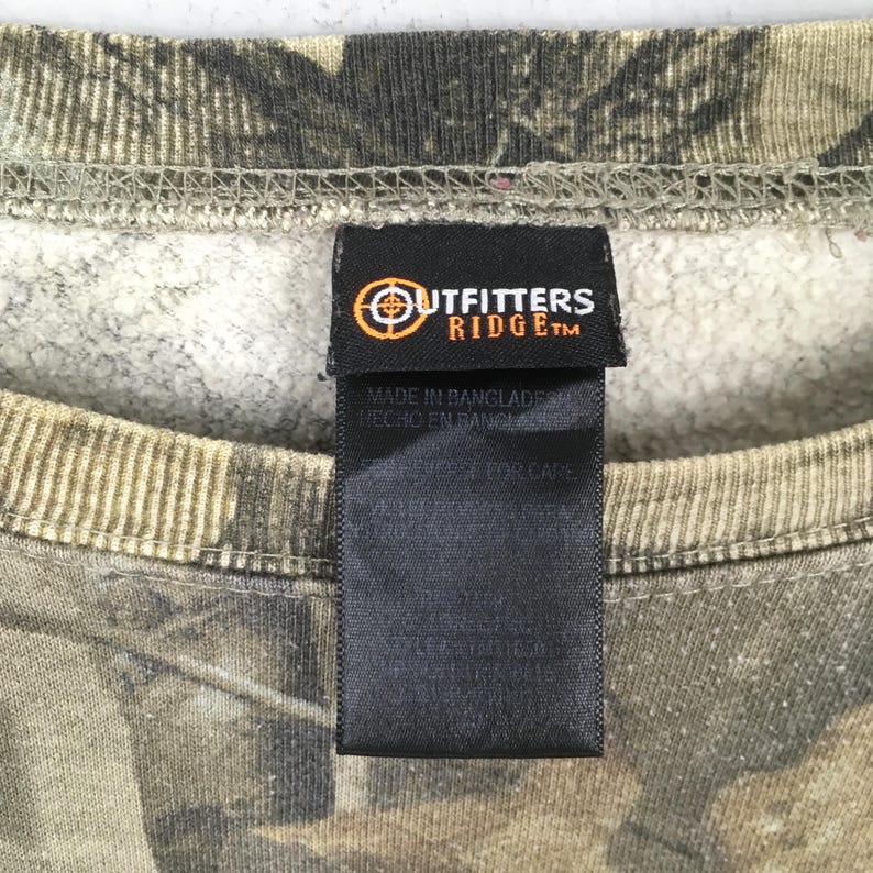 Op de afbeelding: Een sweatshirt met camouflagepatroon, een geribbelde ronde hals en een zwart stoffen label bij de kraag. Het label bevat de tekst Outfitters Ridge met een doellogo, evenals Made in Bangladesh en aanvullende wasvoorschriften.