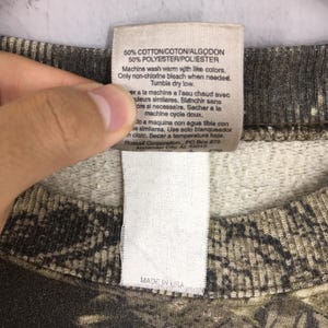 May include: Close-up of a tag from a camouflage-patterned sweatshirt. The tag reads "50% COTTON/COTON/ALGODON, 50% POLYESTER/POLIESTER" and includes washing instructions. The tag also says "MADE IN USA".