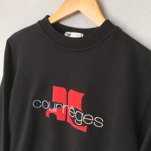 May include: Black sweatshirt with the Courr&egrave;ges logo in red and white embroidery. The logo features a stylized design with the brand name. The sweatshirt has a crew neck and is hanging on a wooden hanger.