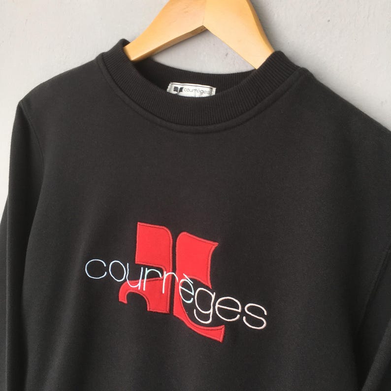 May include: Black sweatshirt with the Courr&egrave;ges logo in red and white embroidery. The logo features a stylized design with the brand name. The sweatshirt has a crew neck and is hanging on a wooden hanger.