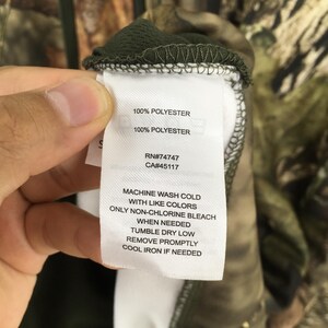 K&ouml;nnte beinhalten: Nahaufnahme eines Etiketts mit Pflegehinweisen und Stoffzusammensetzung. Das Etikett lautet "100% POLYESTER" und "MACHINE WASH COLD WITH LIKE COLORS". Das Etikett enth&auml;lt auch die RN- und CA-Nummern. Das Kleidungsst&uuml;ck hat ein Camouflagemuster.