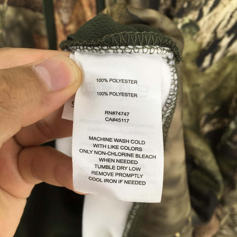 K&ouml;nnte beinhalten: Nahaufnahme eines Etiketts mit Pflegehinweisen und Stoffzusammensetzung. Das Etikett lautet "100% POLYESTER" und "MACHINE WASH COLD WITH LIKE COLORS". Das Etikett enth&auml;lt auch die RN- und CA-Nummern. Das Kleidungsst&uuml;ck hat ein Camouflagemuster.